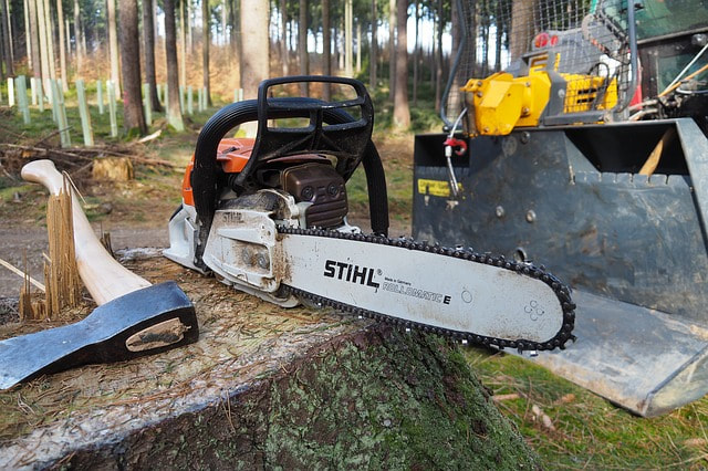 A recently cut log with a chainsaw and ax on it. There is a bulldozer in the background